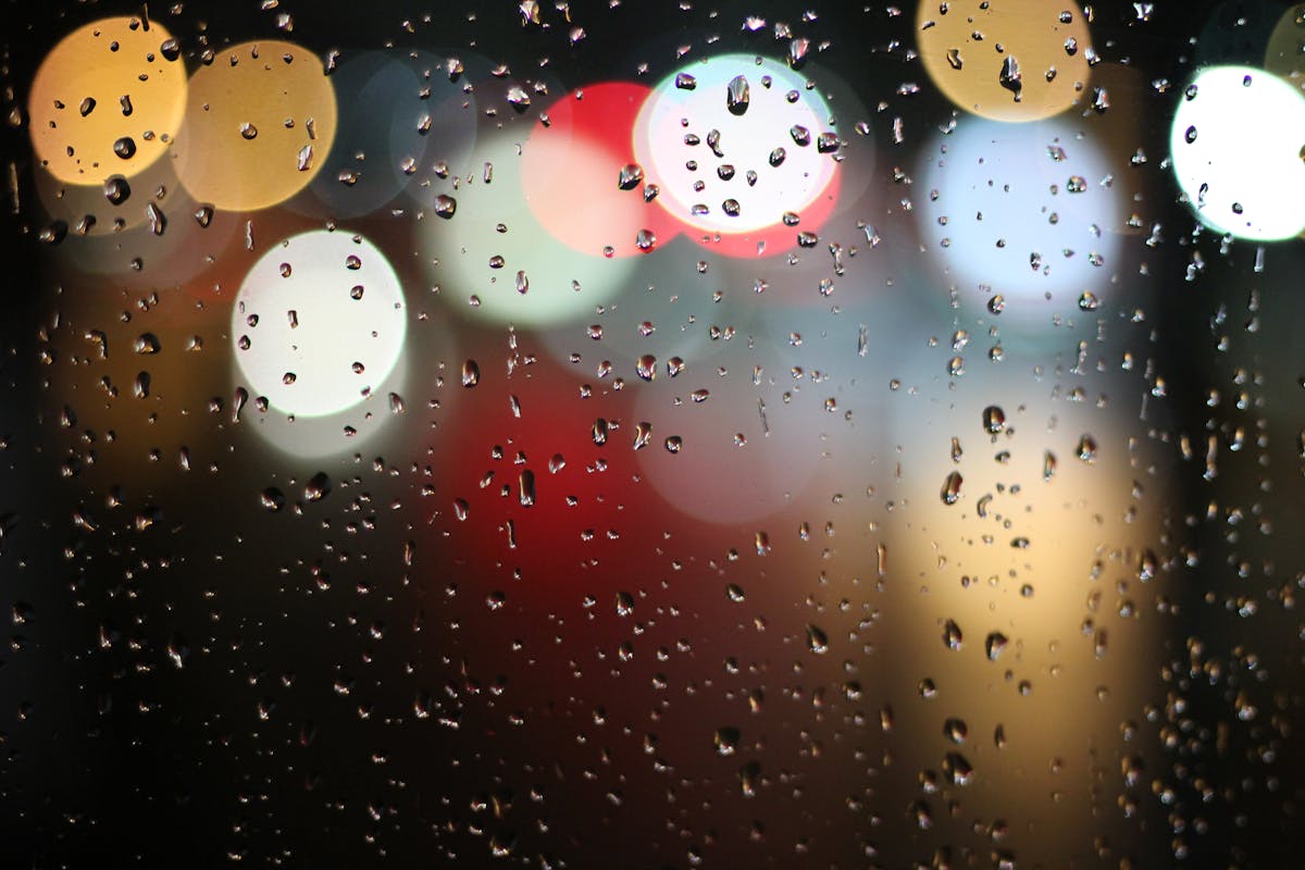 Colorful bokeh lights behind raindrop-covered windowpane, creating a vibrant and moody atmosphere.