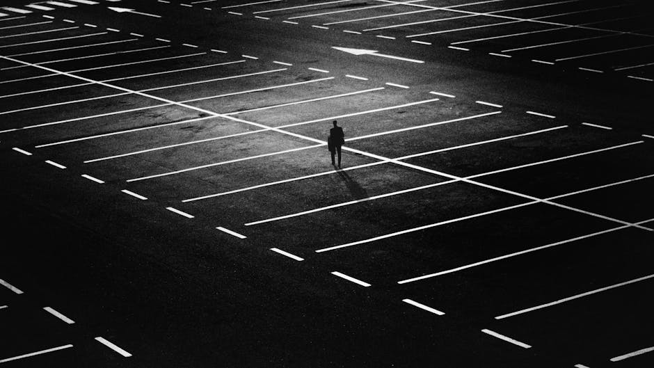 Osoba spacerująca po pustym parkingu, widok z góry, monochromatyczna kompozycja z wyraźnymi liniami miejsc postojowych.