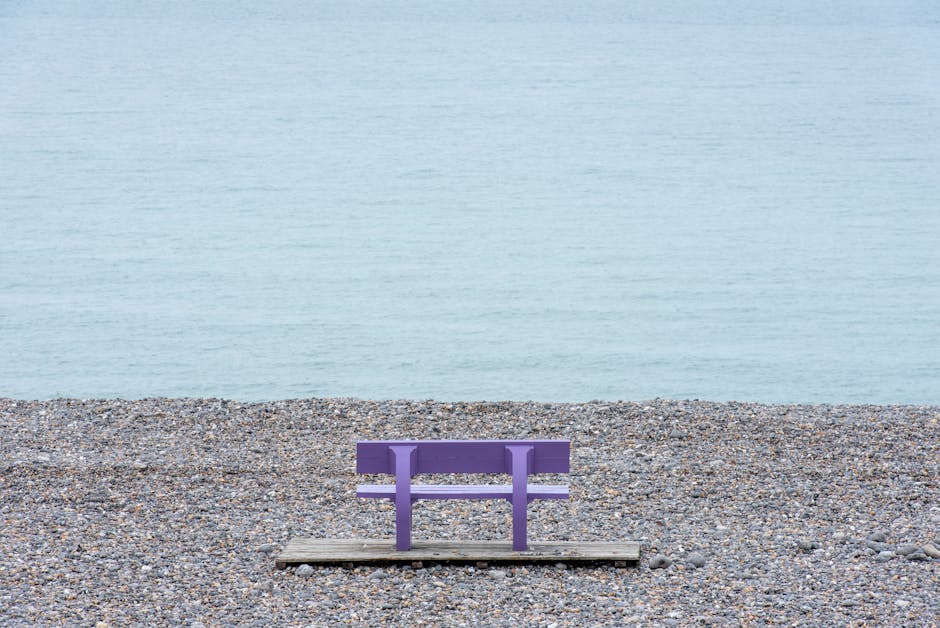 Ławka w kolorze fioletowym na drewnianym podłożu, umieszczona na plaży z kamieniami, z widokiem na spokojne morze.