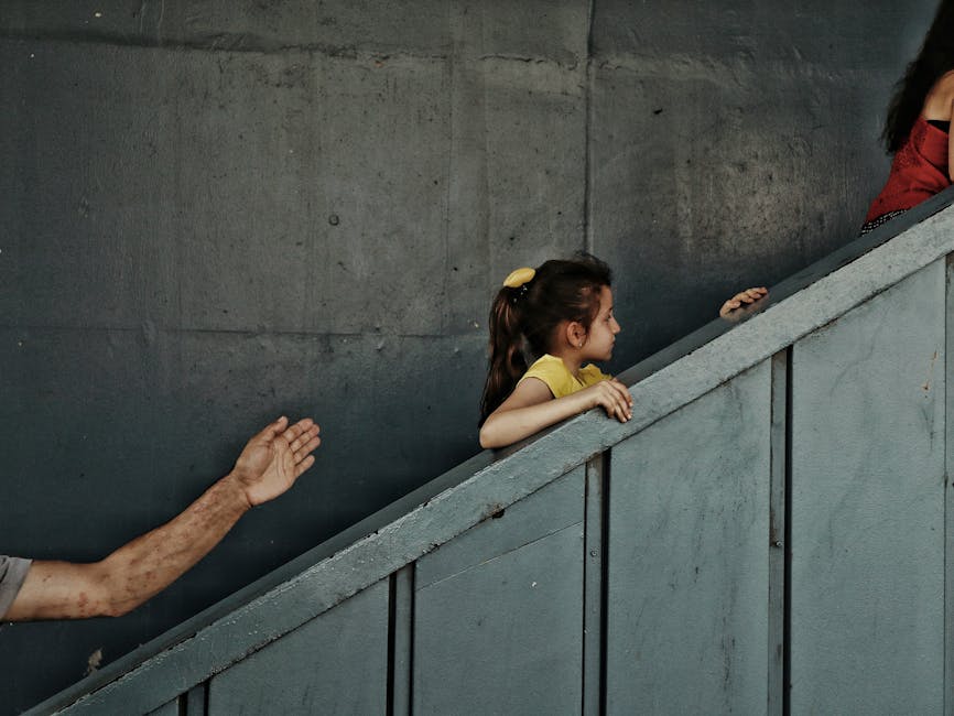 Dziecko w żółtej koszulce wchodzi po schodach, trzymając się poręczy, podczas gdy w tle widać rękę dorosłego. Scena ma kameralny klimat, z szarymi ścianami i naturalnym światłem.