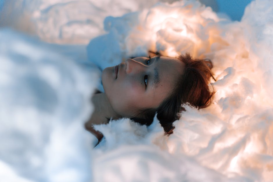 Kobieta leżąca w chmurze białego materiału, z delikatnym oświetleniem w tle, wyrażająca spokój i refleksję.
